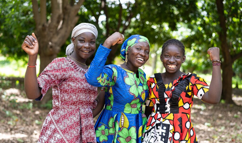 Three African women in colorful traditional dresses raise their fists as a symbol for women’s strength and gender equality.